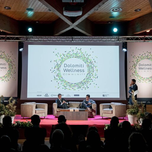 Un evento di wellness nelle Dolomiti, con relatori su un palco attrezzato. Il pubblico assiste mentre viene presentato il tema "Dolomiti Wellness Community".