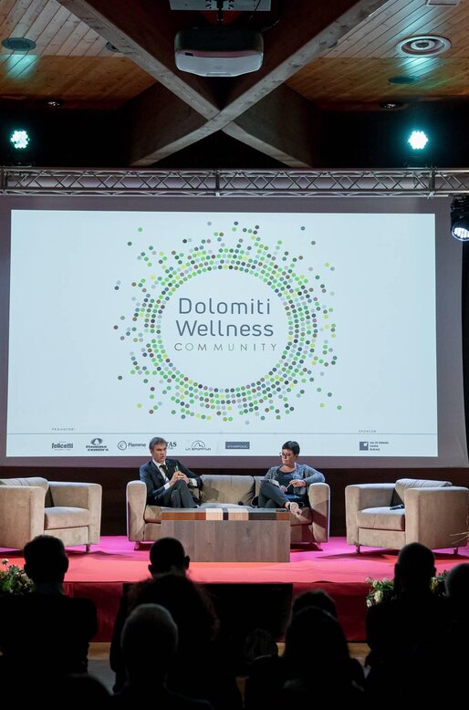 Un evento di wellness nelle Dolomiti, con relatori su un palco attrezzato. Il pubblico assiste mentre viene presentato il tema "Dolomiti Wellness Community".