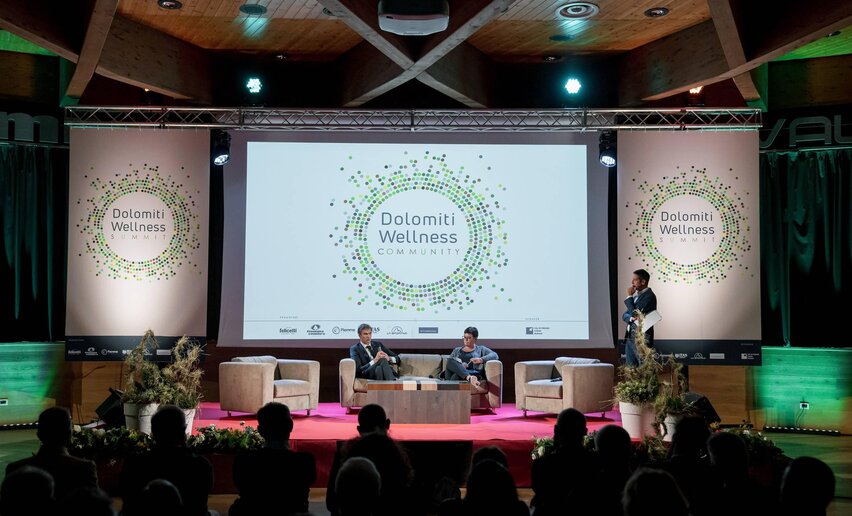 Un evento di wellness nelle Dolomiti, con relatori su un palco attrezzato. Il pubblico assiste mentre viene presentato il tema "Dolomiti Wellness Community".
