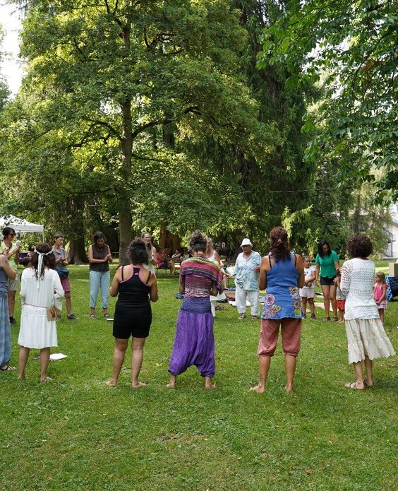 Un gruppo di persone si riunisce in un giardino verde, svolgendo attività all'aperto. Si notano tende bianche e alberi rigogliosi intorno.