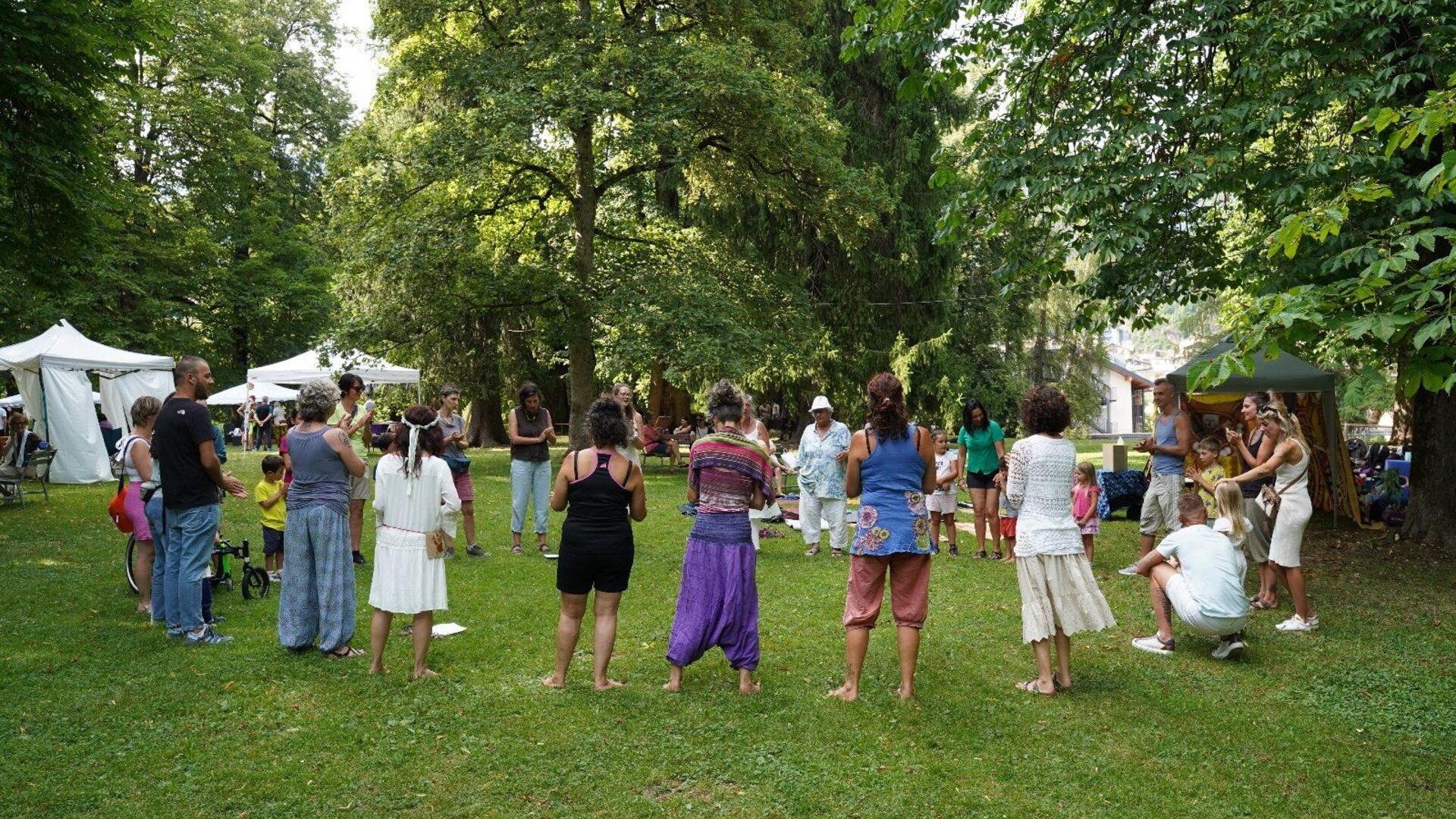 Un gruppo di persone si riunisce in un giardino verde, svolgendo attività all'aperto. Si notano tende bianche e alberi rigogliosi intorno.
