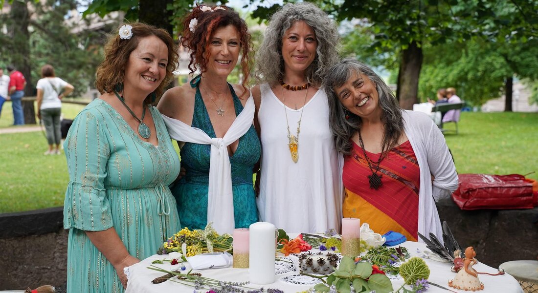 Quattro donne sorridenti si trovano accanto a un tavolo decorato con erbe e fiori. Sullo sfondo si vede un parco verde con altre persone.