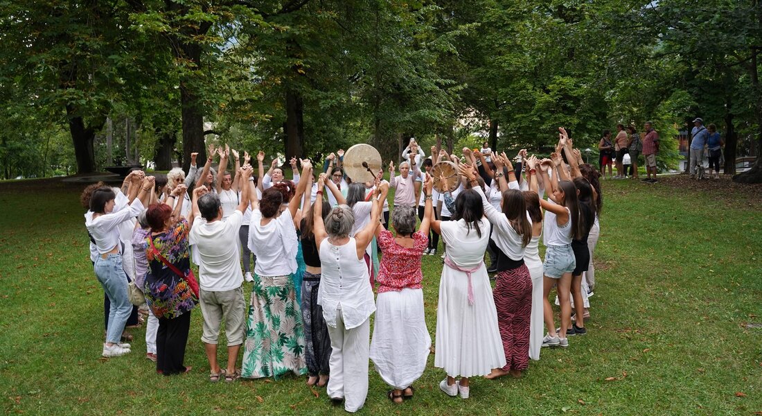 Un gruppo di persone si raduna in un parco, sollevando le mani verso il cielo. Indossano abiti bianchi e colorati, creando un'atmosfera di festa e comunità.