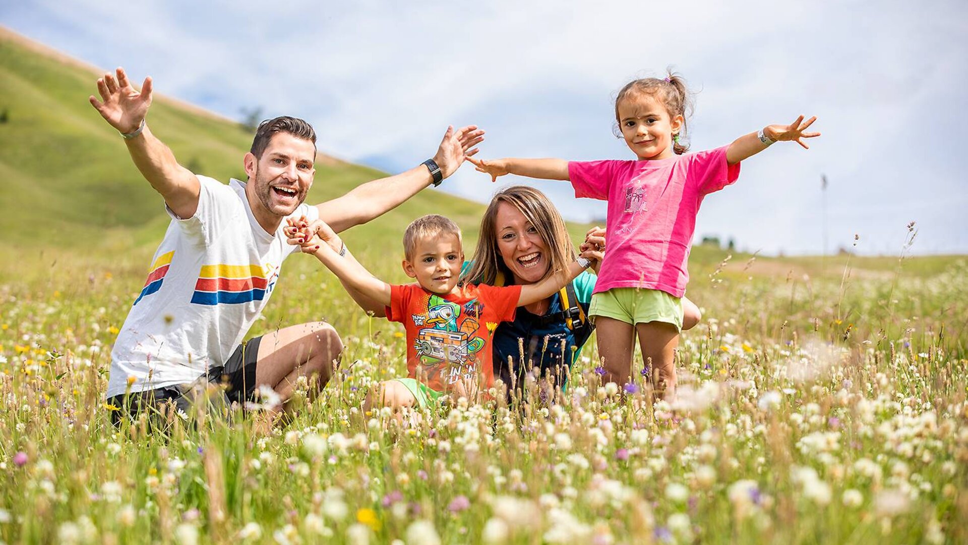 Genitori con bambini in un prato fiorito alzano le braccia al cielo contenti | © APT Fiemme e Cembra