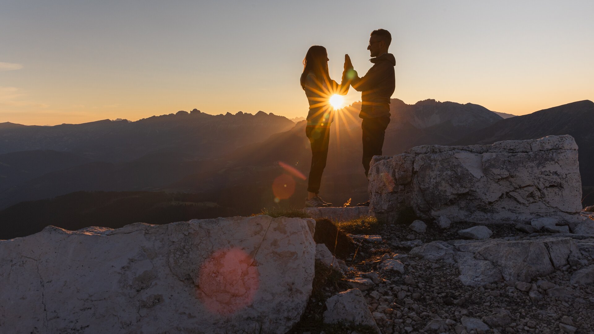 Coppia all'alba in montagna; i raggi del sole passano tra le loro mani giunte | © APT Fiemme e Cembra - foto di Gaia Panozzo