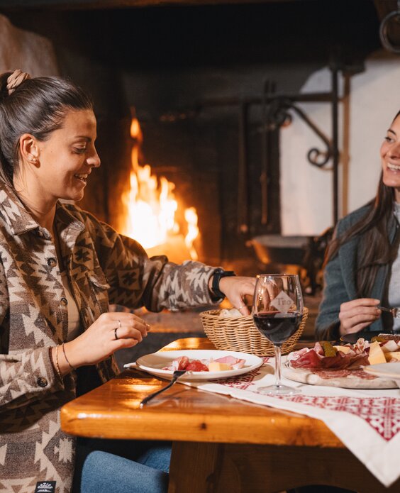Due amiche condividono un tagliere di salumi e delle castagne, accompagnando il tutto con un bicchiere di vino rosso | © APT Fiemme e Cembra - foto di Gaia Panozzo